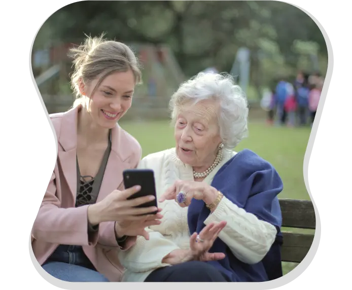 Eine junge Dame zeigt einer älteren Frau etwas auf ihrem Smartphone. Sie sitzen auf einer Bank im Grünen.